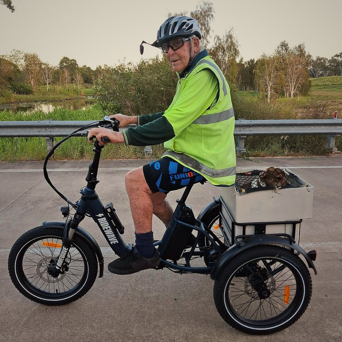 A rider on a black Wallaby electric trike on a riverside path, his dog in the rear box