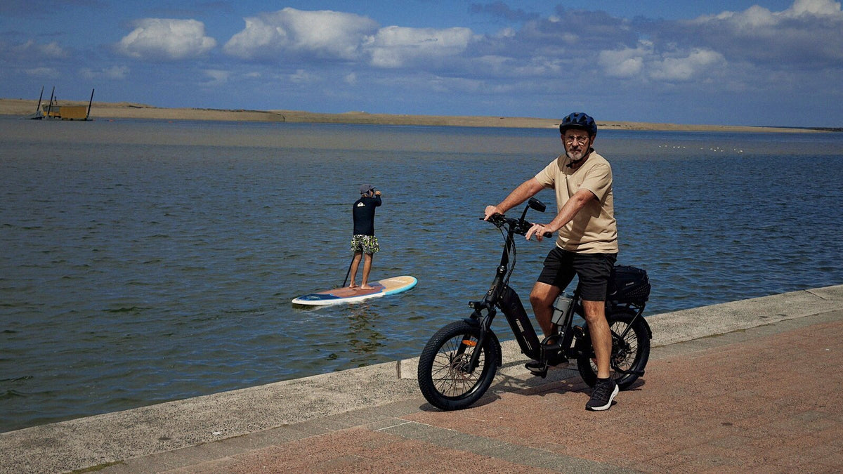 A rider on a Ridewave e-bike on a waterfront path while a paddleboarder glides past