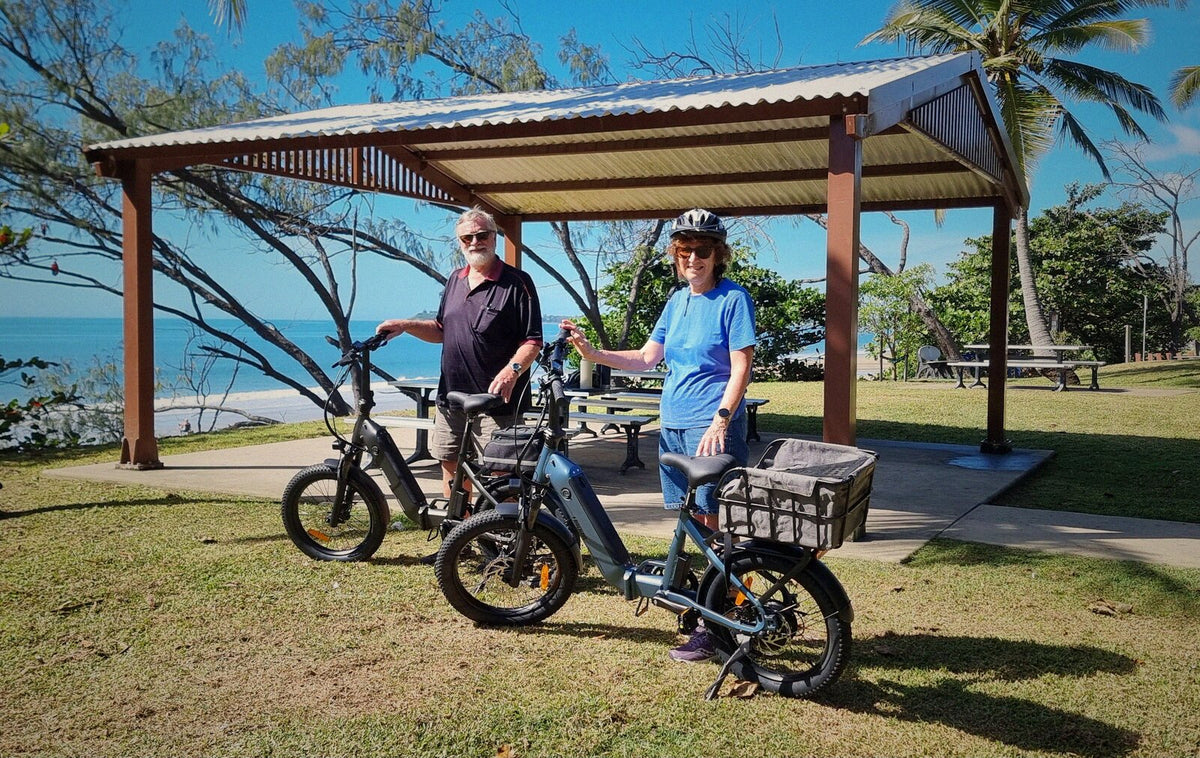 Two riders with Ridewave Miniwave e-bikes under a seaside picnic shelter